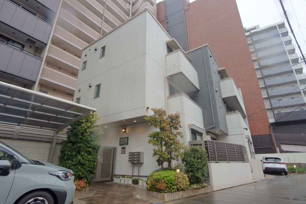 メゾン・ド・メトロ西中島南方のオフホワイトとグレイの綺麗な3階縦集合住宅の外観。エントランス前はいくつかの花木が植樹されていて緑が映えている。